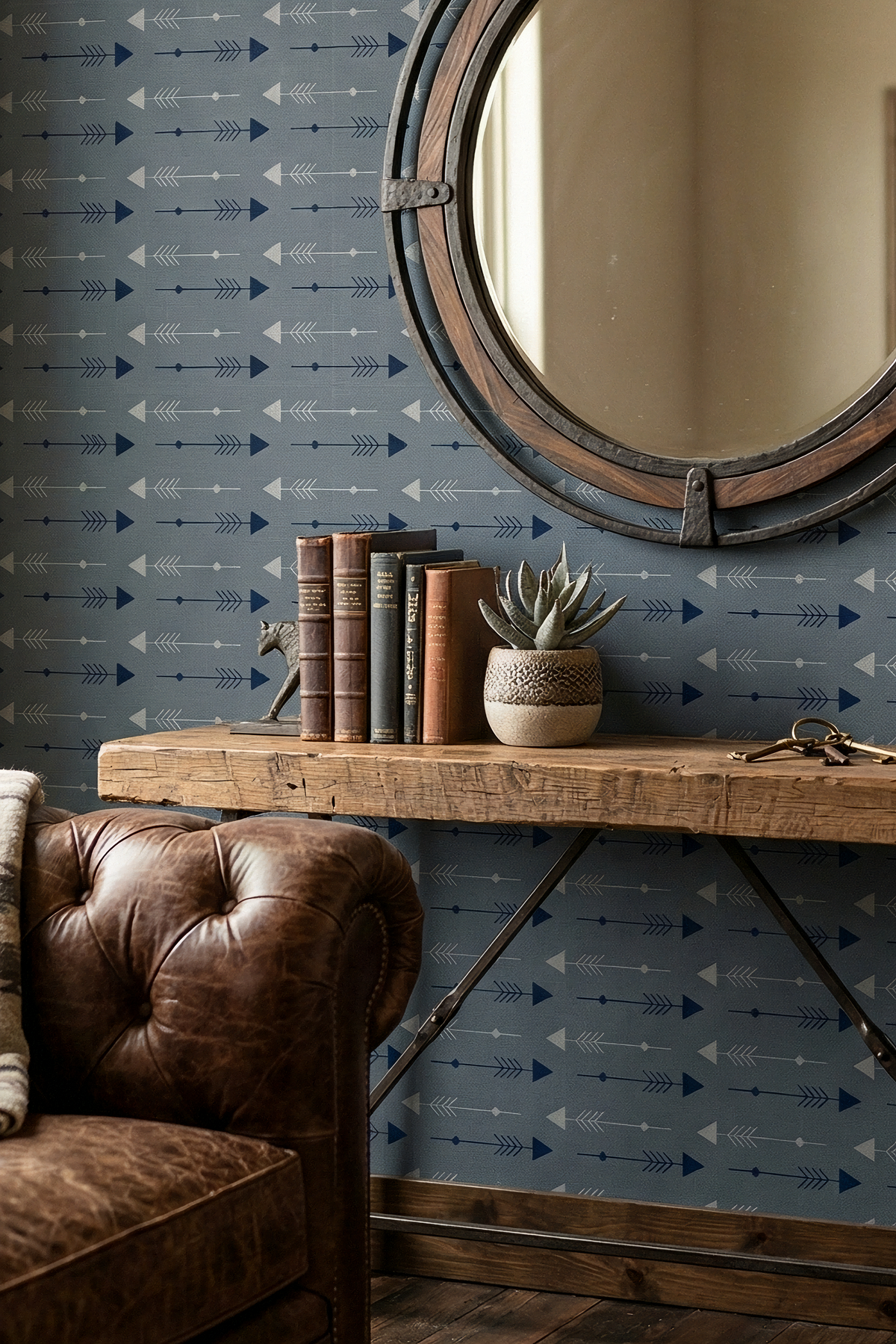 A styled interior scene featuring a rustic wooden console table against a blue wallpaper with a repeating white and navy arrow pattern. A round wooden-framed mirror hangs on the wall, next to a brown leather tufted armchair.