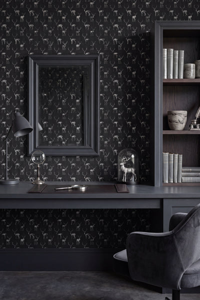 A styled interior scene featuring dark charcoal wallpaper with a repeating deer pattern, a matching dark desk, a large framed mirror, a bookshelf, and a gray chair.