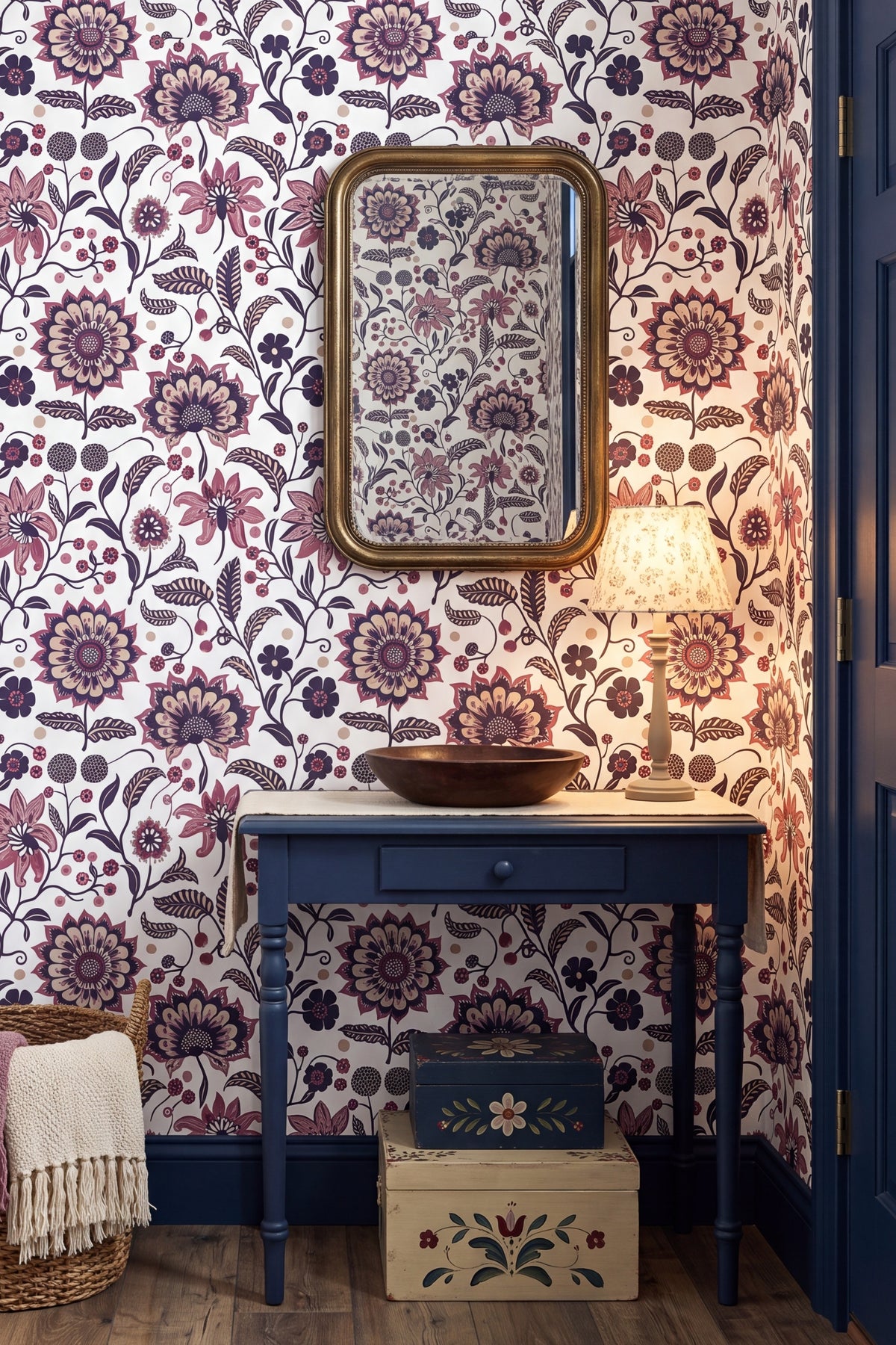 A styled interior corner featuring floral wallpaper in purple, pink, and brown tones, a gold-framed mirror, a blue console table with a bowl sink, a lit table lamp, and decorative storage boxes on the floor.