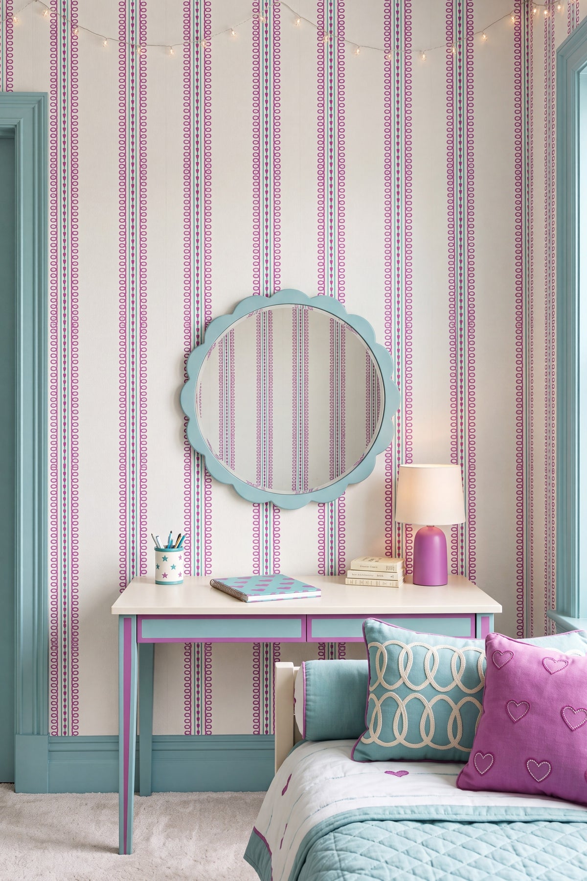 A styled bedroom corner featuring pink and white vertically striped wallpaper, a light blue scalloped mirror, a matching light blue and pink writing desk, a pink lamp, and a bed with patterned pillows.