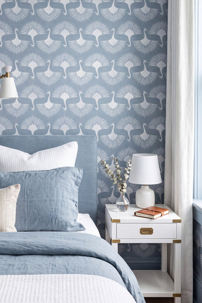 A styled bedroom corner featuring a blue and white patterned wallpaper with a repeating bird motif, a bed with blue bedding, a white nightstand, and a window with sheer curtains.