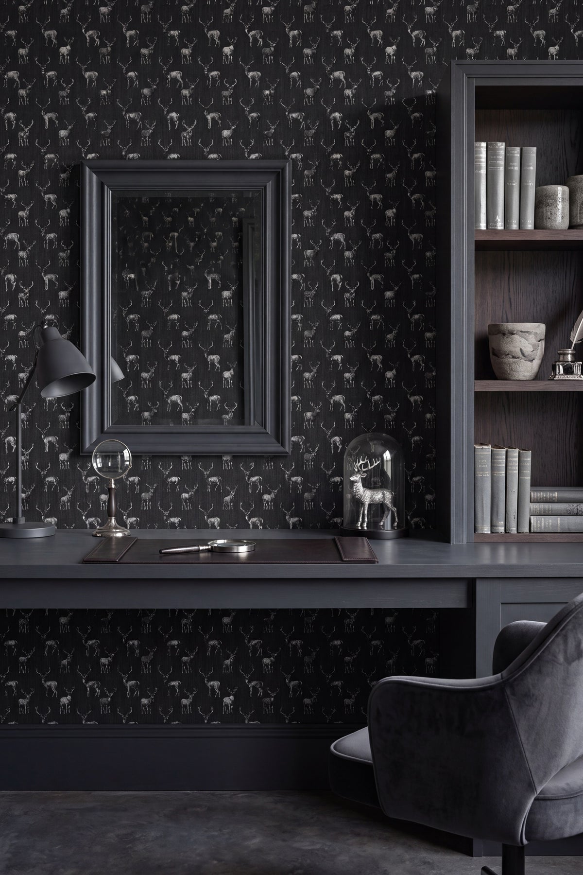 A styled interior scene featuring dark charcoal wallpaper with a repeating deer pattern, a matching dark desk, a large framed mirror, a bookshelf, and a gray chair.
